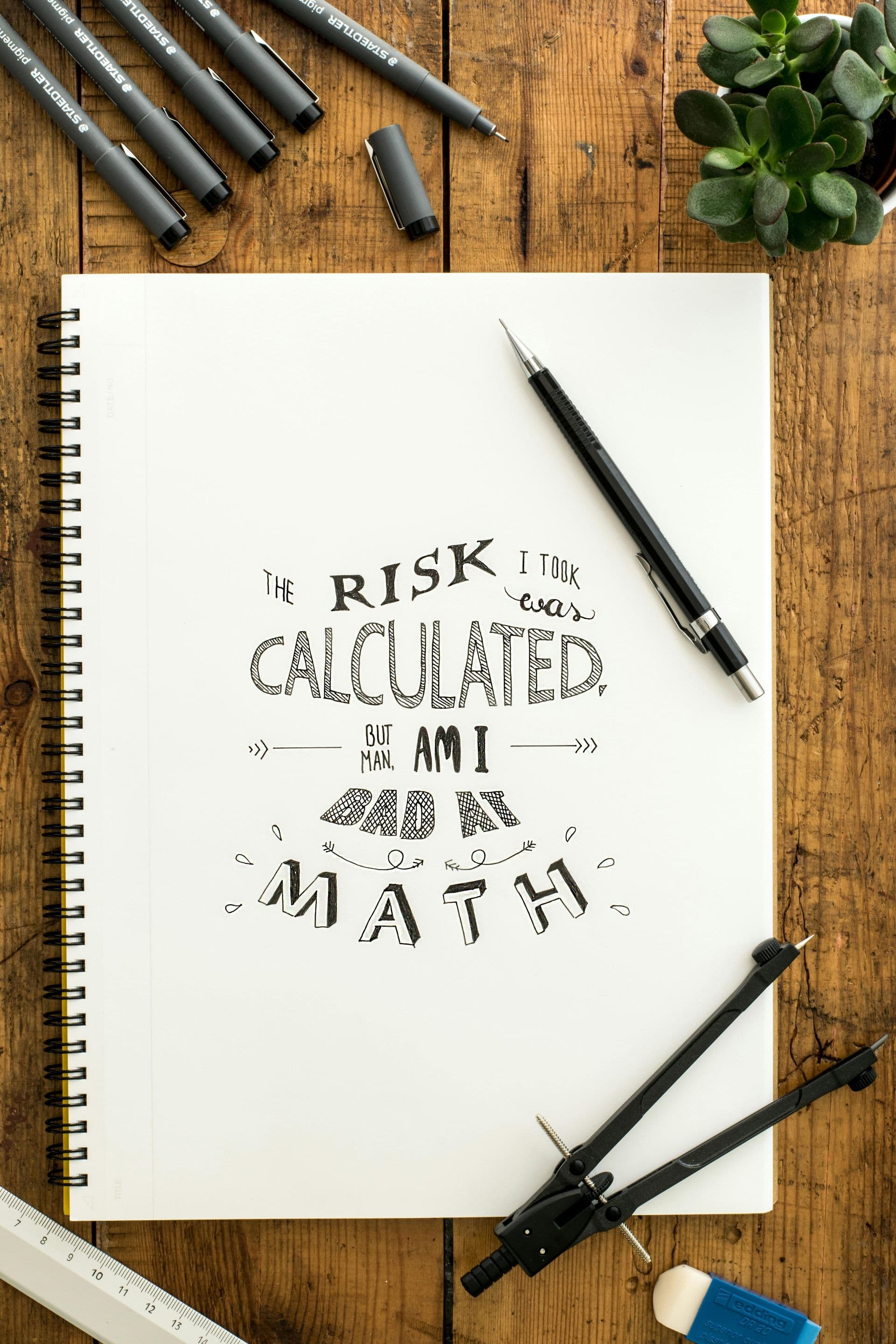 A sketchbook lettered with "The risk I took was calculated, but man, am I bad at math", surrounded by pens and drafting tools.